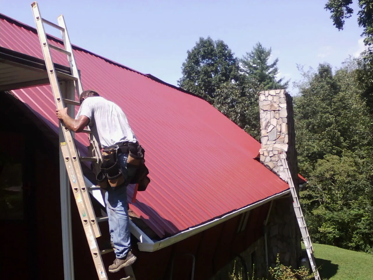 Roofer on a ladder beside a red metal roof and stone chimney — Soffit & Fascia Repair by Roofing Brothers in Marcus Hook